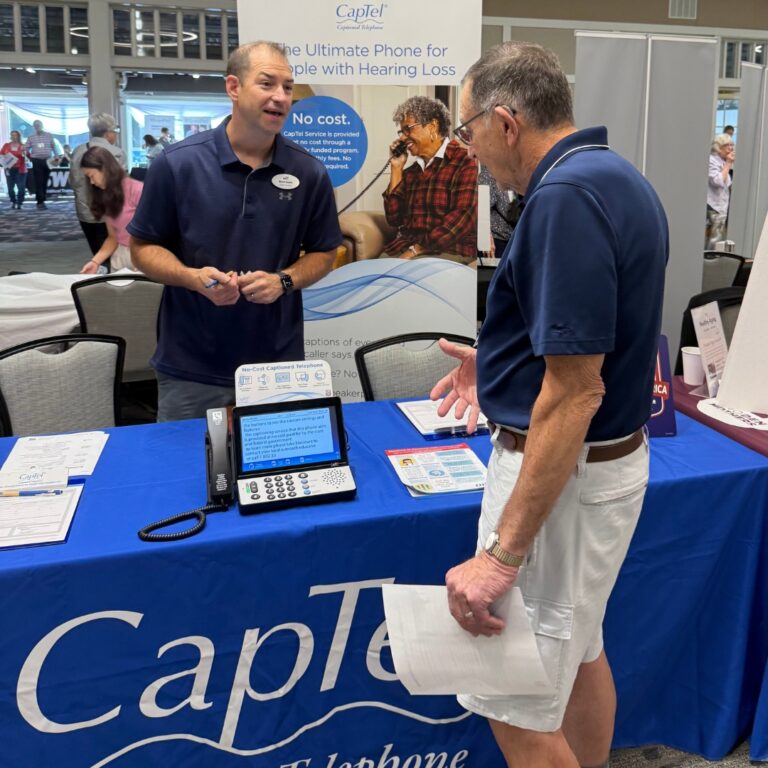 Two people talking over an exhibitor table, a landline phone with a screen sits on the table.