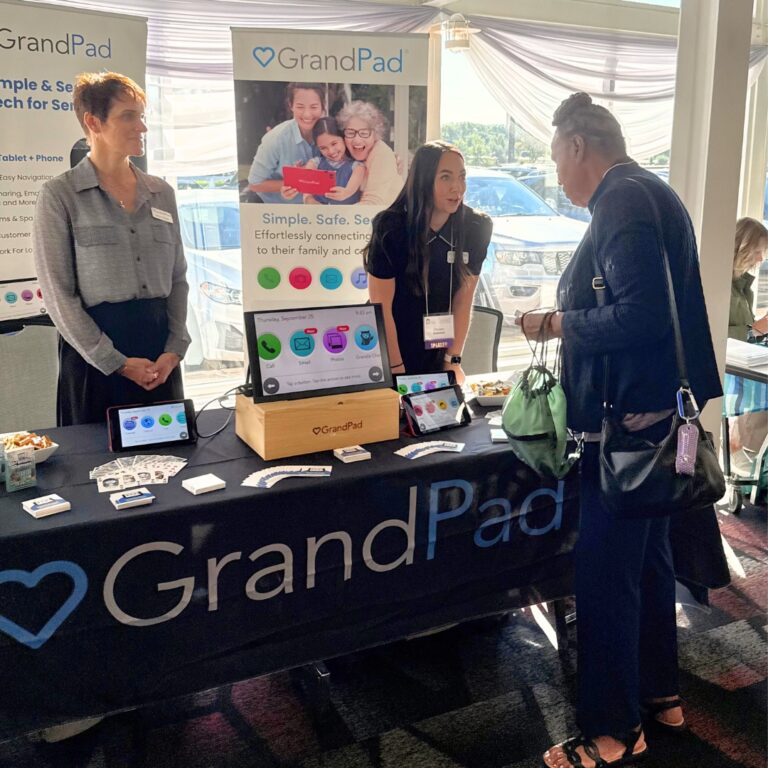 Two people talking to a woman at an exhibitor table, a tablet with a large screen sits on the table.